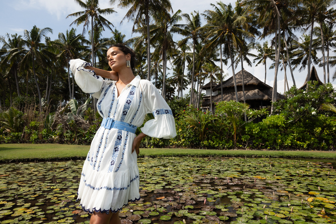 Mannequin portant une robe blanche brodée bleu avec ceinture bleue, posant devant un étang aux nénuphars dans un jardin tropical avec palmiers.
